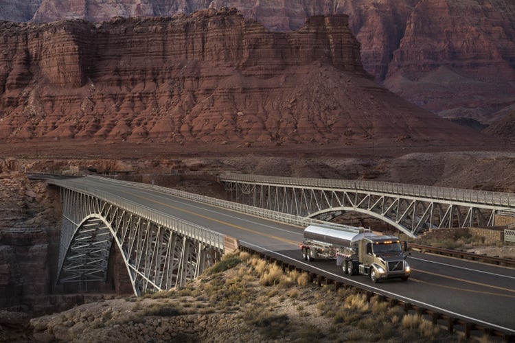 A Volvo VNR truck with a fuel tanker crosses a steel bridge in a desert canyon landscape.